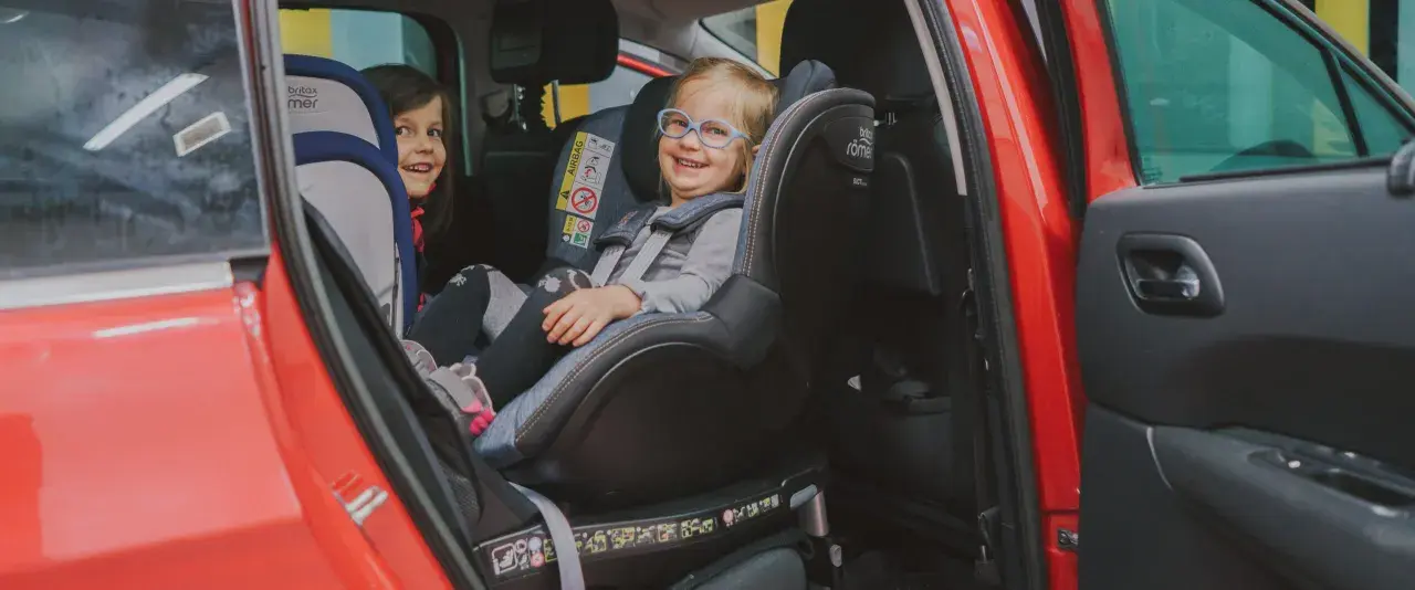Dwie uśmiechnięte dziewczynki w fotelikach Britax Romer 0-18 kg w czerwonym samochodzie.
