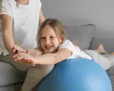 Dziecko z obniżonym napięciem mięśniowym ćwiczy mowę, leżąc na piłce rehabilitacyjnej pod opieką terapeuty.