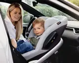 Uśmiechnięta mama patrzy na dziecko w foteliku z przodu. Widoczna jest poduszka powietrzna pasażera.