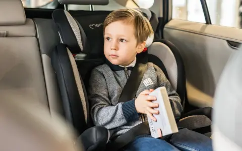 Chłopiec w foteliku samochodowym (testy fotelików 9-18 kg) trzyma książkę, gotowy na podróż.