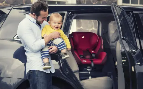 Tata z uśmiechniętym dzieckiem przy samochodzie, obok czerwonego fotelika Joie i-Spin 360.