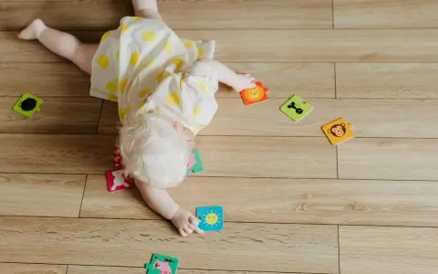 Dziecko w żółtej sukience z kółkami próbuje wykonać obrót z brzucha na plecy, otoczone kolorowymi puzzlami ze zwierzątkami.