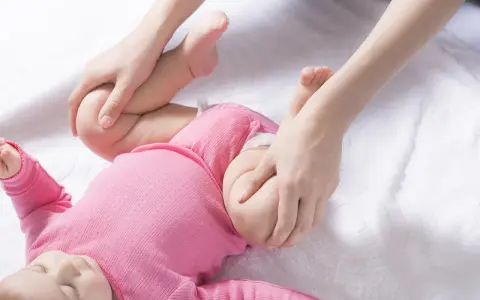 Delikatne ruchy dłoni pomagają w diagnostyce dysplazji stawu biodrowego u niemowlaka w różowym body.