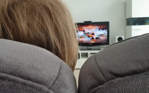 Niemowlę z blond włosami ogląda bajkę na telewizorze w tle, siedząc na kanapie.