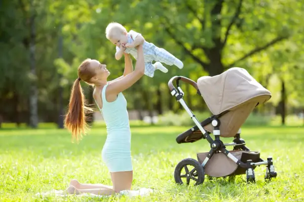 Uśmiechnięta mama podrzuca dziecko w powietrze na trawie obok wózka.