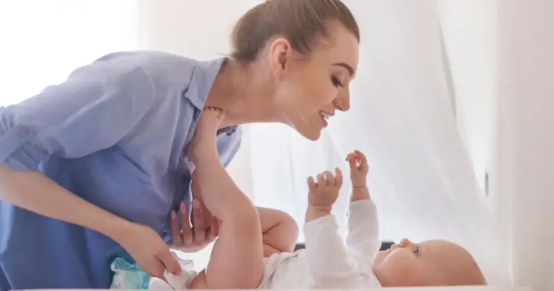 Uśmiechnięta mama zmienia pieluchę dziecku, zapewniając mu komfortowe szerokie pieluchowanie bioderek.